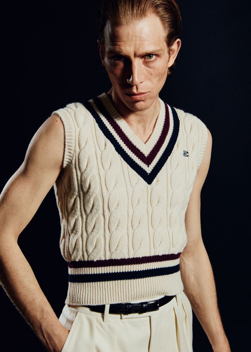 An adult male poses indoors, wearing a cable-knit sweater vest with a V-neck, off-white pleated trousers, and a dark belt against a dark backdrop.