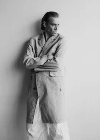 Black and white portrait of a tall, slender man in an open beige coat, standing with arms crossed against a neutral wall. 