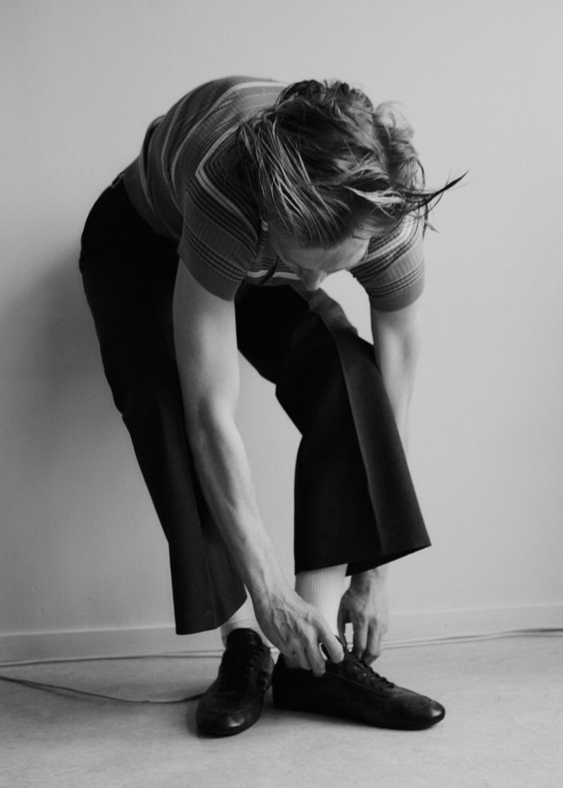 Black and white photo of a person bending down to tie their shoes, wearing a striped shirt and dark pants, against a neutral wall.