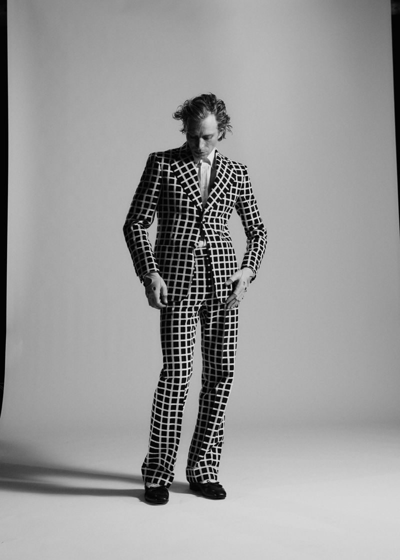 A man in a black-and-white checkered suit poses in a photo studio. The shot captures his full body against a neutral backdrop.