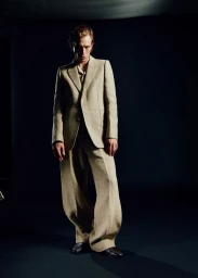 Model Mike Vasilakis wearing a tailored beige suit with wide-leg trousers against a dark seamless studio backdrop.