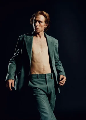 Model Mike Vasilakis wearing an open teal tailored suit jacket and matching trousers without a shirt against a dark background.