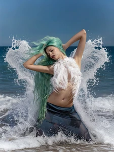 Woman with long mint green hair posing in seawater with dramatic white water splashes framing her body against an ocean backdrop