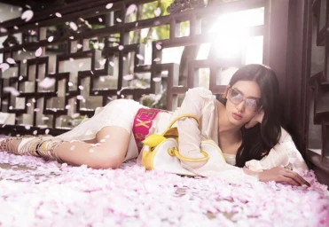 Model Ying Ouyang wearing tinted sunglasses and a white dress reclining on pink flower petals in front of a wooden lattice screen.