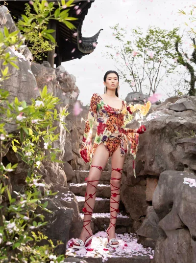 Model Ying Ouyang poses in a floral printed draped top and high red lace-up sandals on rocky garden steps with falling petals.