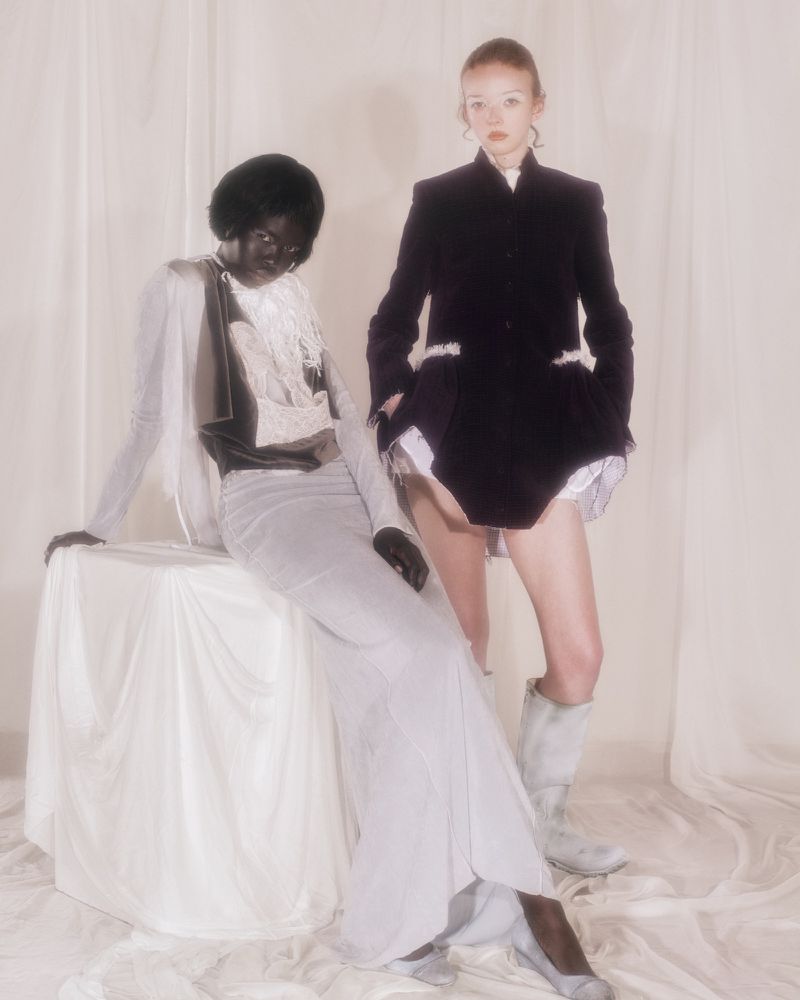 Lily Nova and a model posing in draped and tailored garments against a cream fabric backdrop.