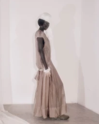 A Black female model stands in profile wearing a sheer taupe floor-length dress against a neutral white studio wall.