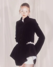 Model Lily Nova posing in a black structured high-collar jacket with flared peplum against a pale draped fabric backdrop.