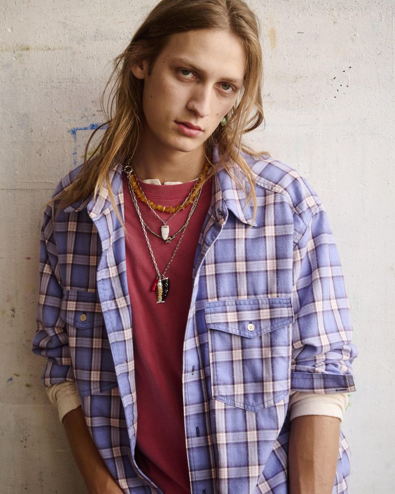 Young man with long hair wearing an open purple plaid shirt over a red t-shirt with layered necklaces against a textured wall.