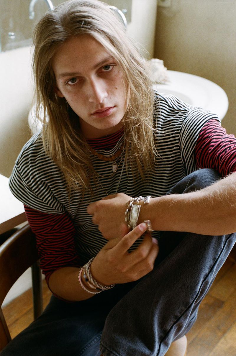 Model with long blonde hair wearing layered striped t-shirts and silver bracelets, seated on a chair in a bathroom.