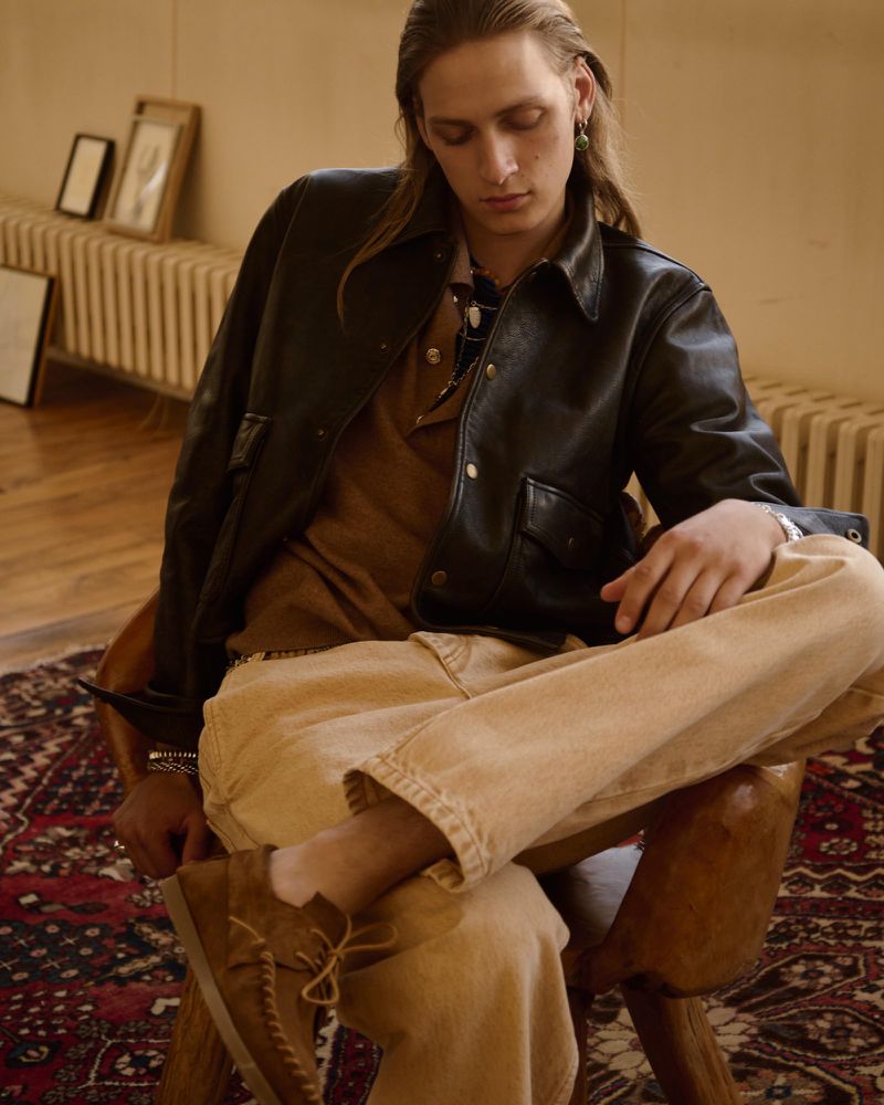 Model Jesse Rinderknecht seated in a wooden chair wearing a black leather jacket, brown top, and beige trousers on an ornate rug.