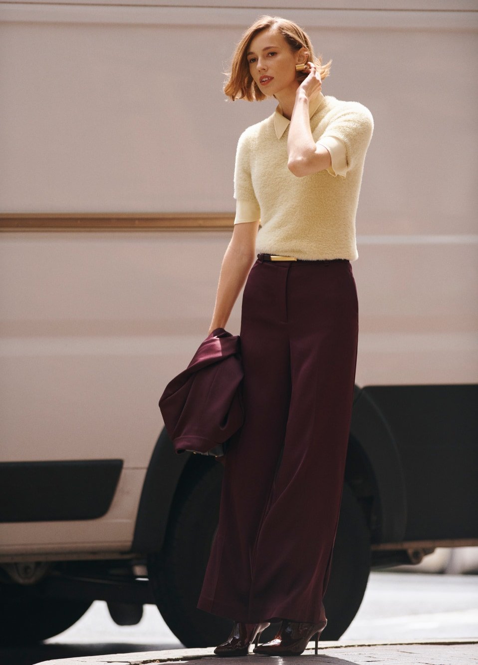 A stylish adult female in fashionable outerwear with high heels poses against a neutral van, offering a modern, urban aesthetic.
