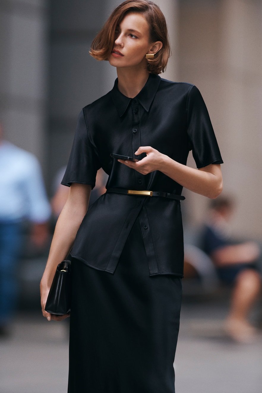Fashionable woman in black shirt dress on city street holds a smartphone and purse, showcasing modern urban style and elegance.