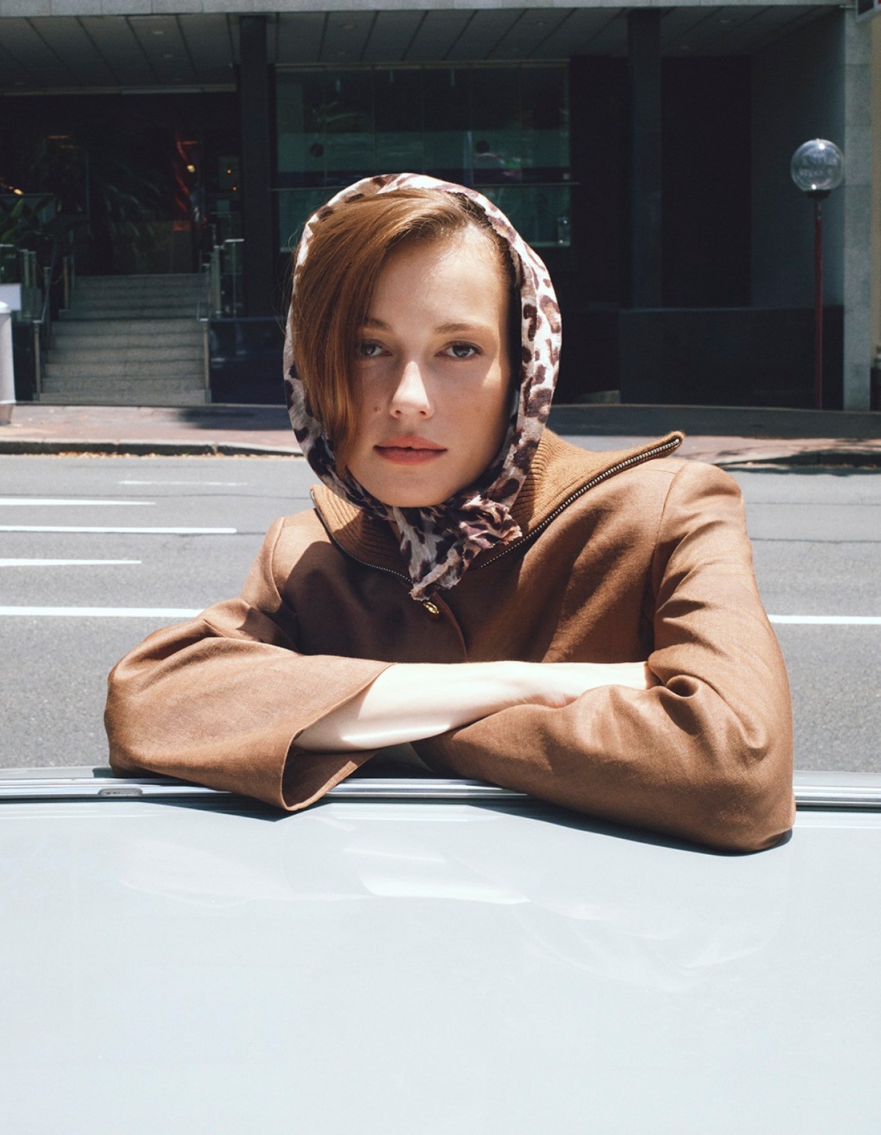 Outdoor portrait of an adult female with red hair wearing a headscarf and brown jacket, posing in an urban setting in natural light.