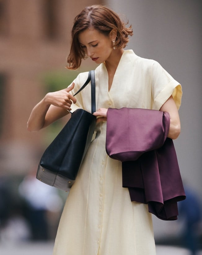 Woman in cream dress opening black handbag while holding a plum jacket outdoors.