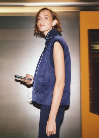 A young adult woman in a navy vest holds a smartphone, set against a neutral indoor background with a hint of orange, creating a modern, casual vibe.