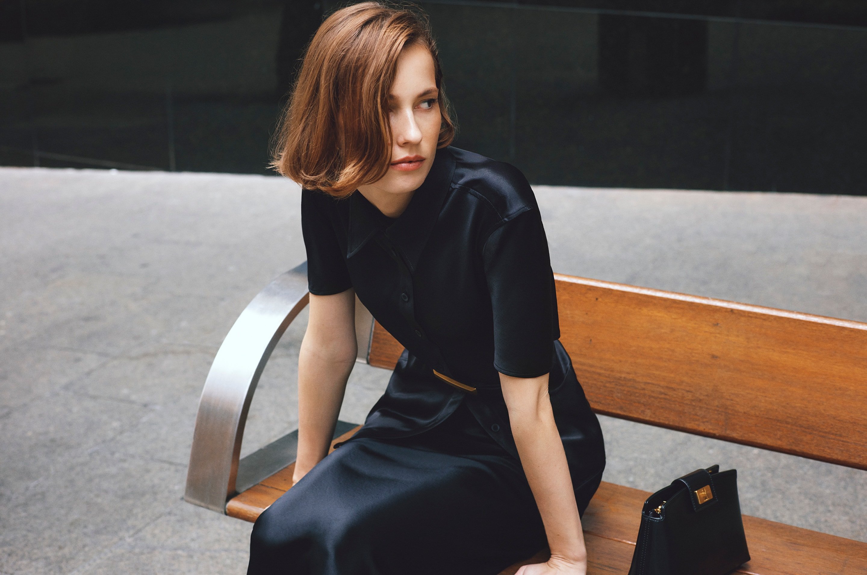 Woman with short brown hair wearing a black silk dress seated on a wooden outdoor bench for Country Road.