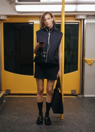 Woman standing in a subway car using her smartphone, wearing a black vest, dress, socks and shoes, and holding a tote bag.
