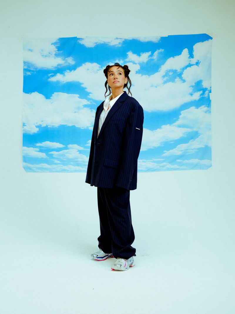 Full-length portrait of a woman wearing an oversized pinstripe suit and athletic shoes against a blue sky and cloud backdrop.