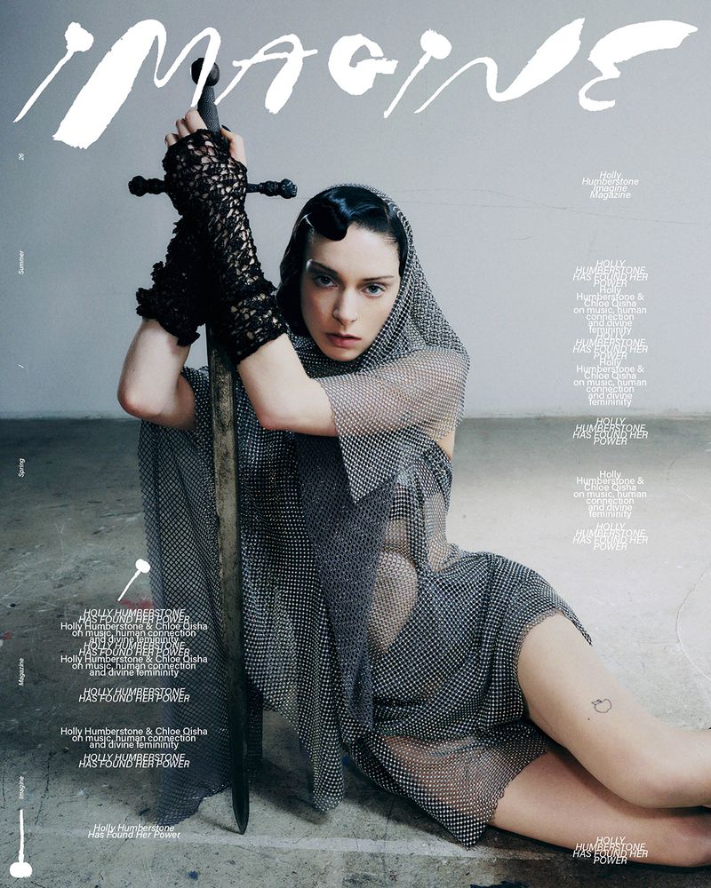 Holly Humberstone wearing a silver chainmail hooded dress and black lace cuffs while holding an upright sword.