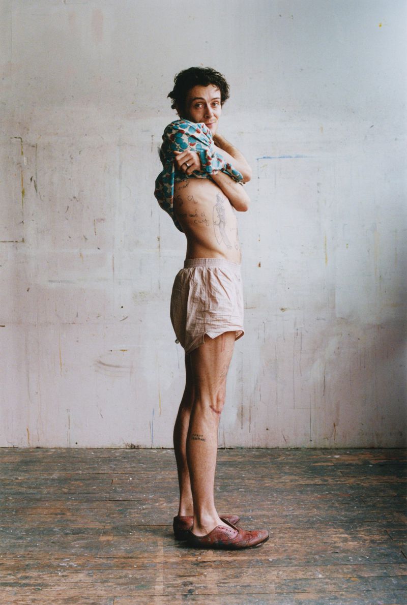 Model Sonny Hall pulling up a printed top while wearing light pink shorts against a distressed studio wall.
