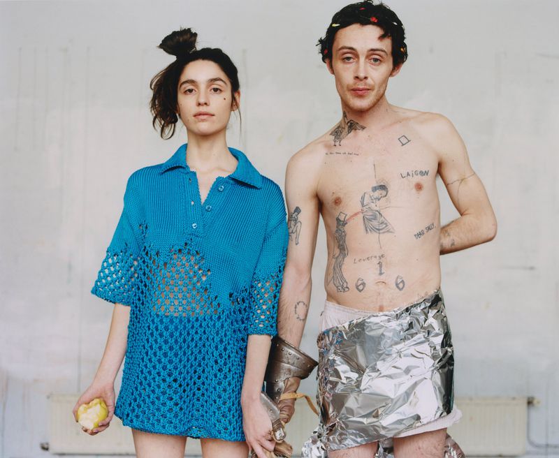 Model Sonny Hall shirtless in silver foil wrap standing next to a woman in a bright blue knit polo dress against a pale wall.