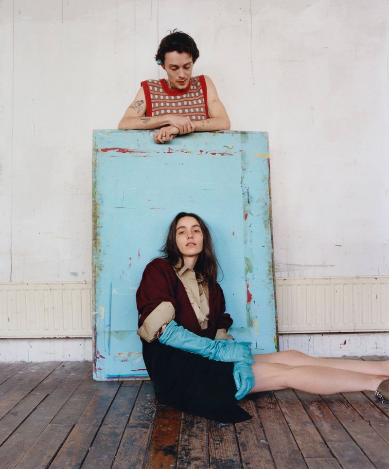 Two models posed against a large painted blue panel, featuring a man in a red patterned tank top and a woman in long blue gloves.