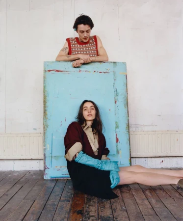 Two models posed against a large painted blue panel, featuring a man in a red patterned tank top and a woman in long blue gloves.