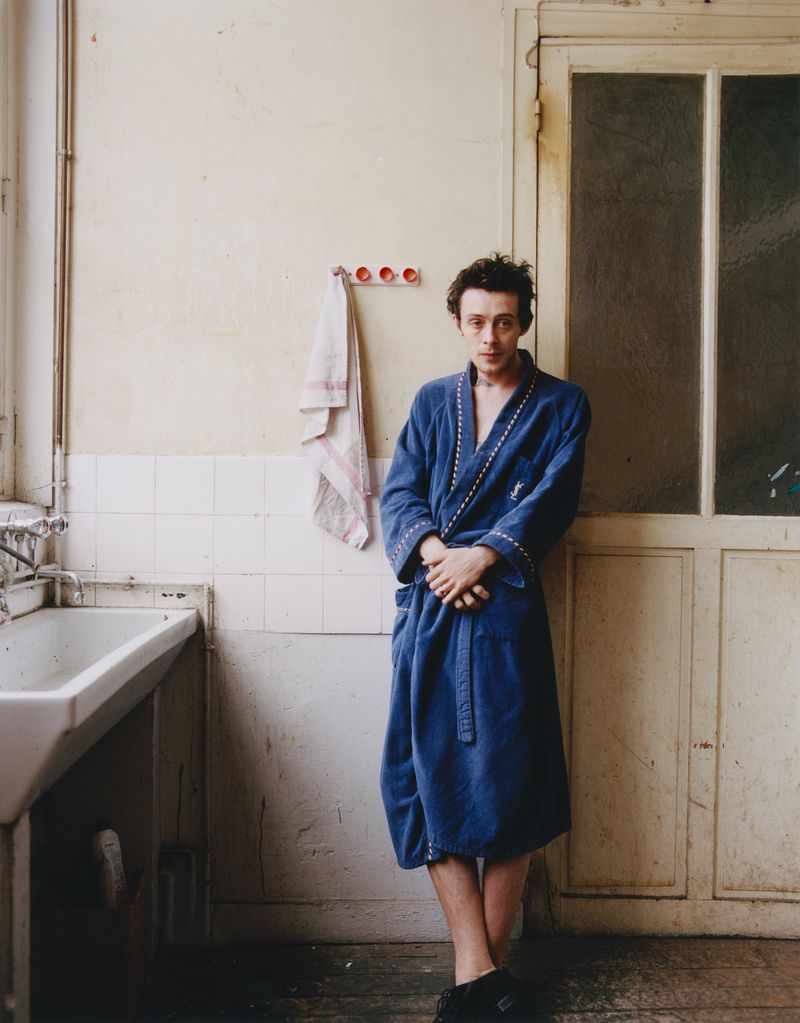 Sonny Hall standing in a blue belted bathrobe next to a deep sink in a rustic bathroom with aged walls and a wooden door.