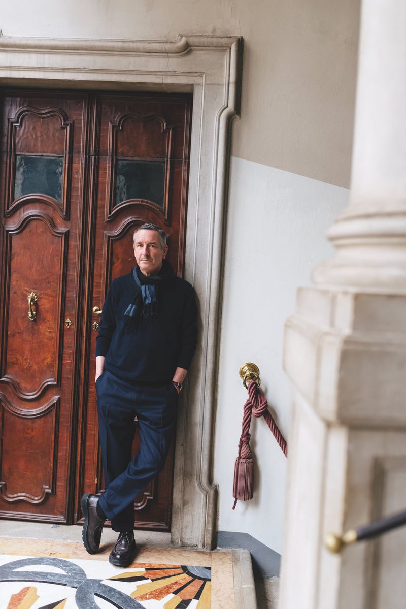 Fashion designer Dries Van Noten wearing a navy sweater and trousers, leaning against an ornate wooden doorway.