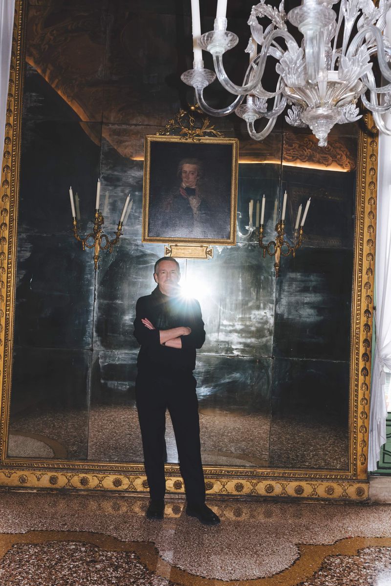 Dries Van Noten standing with arms crossed in front of a large antique gilded mirror and ornate chandelier.