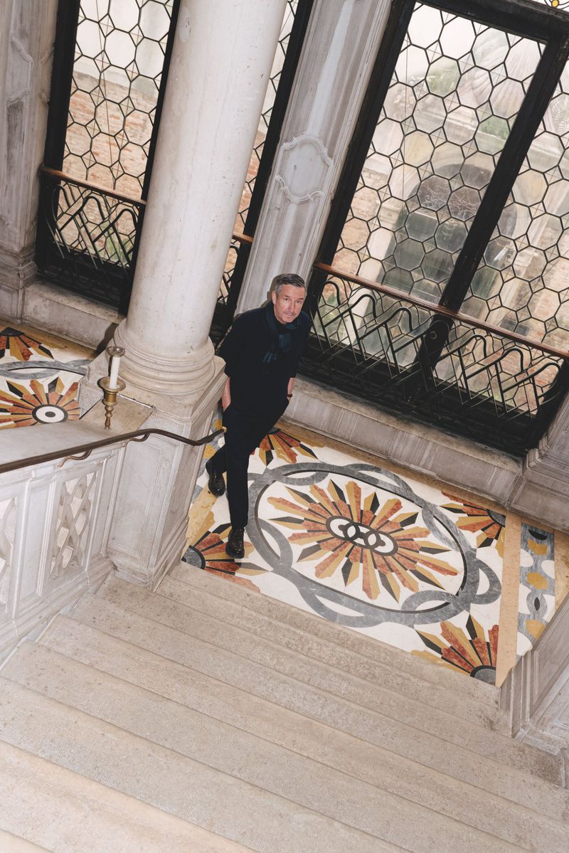 Fashion designer Dries Van Noten in black standing on a grand marble staircase landing with mosaic flooring.