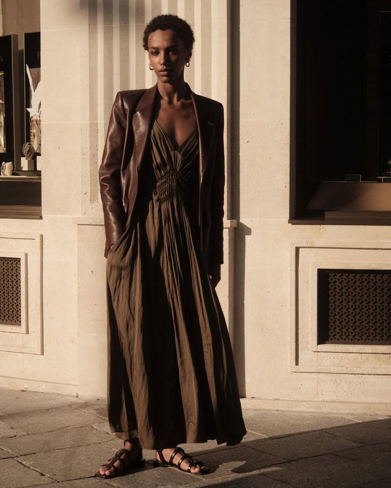 Model Saadia Bodenmann wearing a pleated olive maxi dress and brown leather blazer standing outside in warm sunlight.