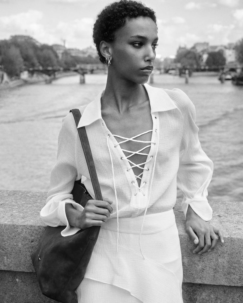 Model Saadia Bodenmann in a white lace-up blouse and shoulder bag leaning on a stone bridge in a black and white fashion portrait.