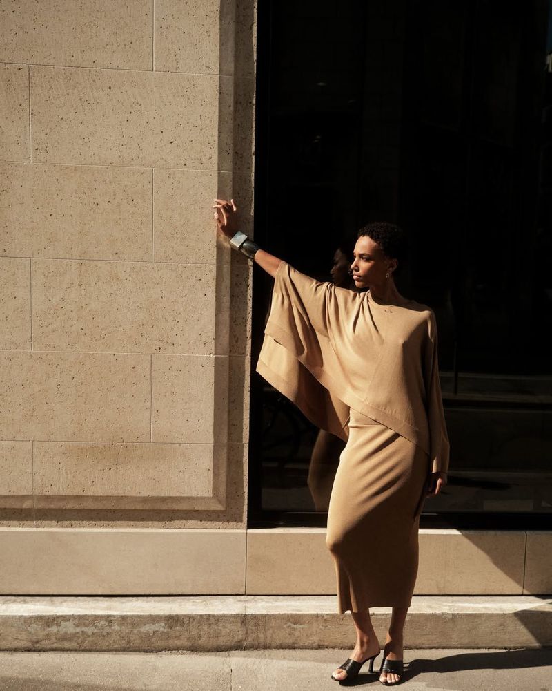 Model Saadia Bodenmann wearing a beige draped knit dress and black heels against a stone wall for Barbara Bui.
