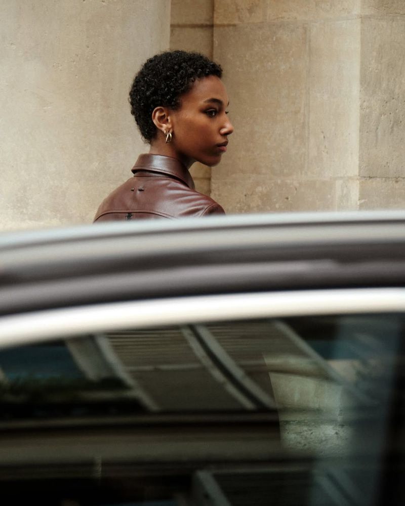 Profile of a woman with short natural hair wearing a brown leather jacket viewed over a car roof against a stone building wall.