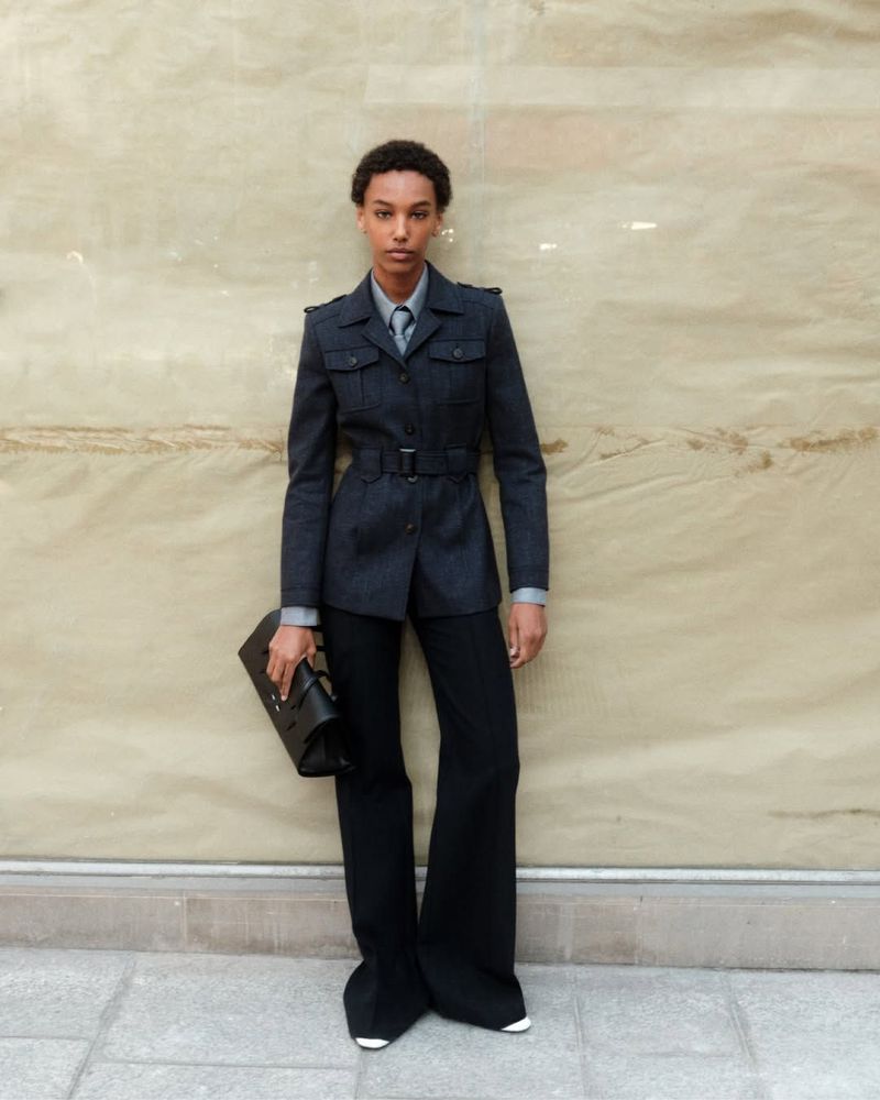Model standing outdoors wearing a dark belted utility jacket and trousers while holding a black clutch bag.