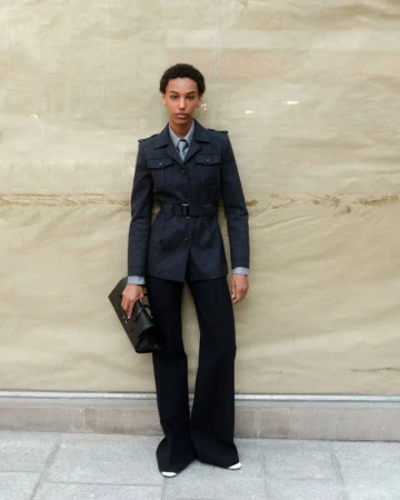 Model standing outdoors wearing a dark belted utility jacket and trousers while holding a black clutch bag.