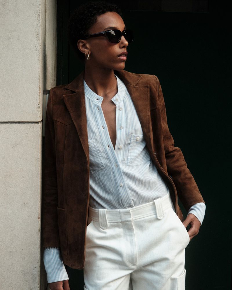 Model in sunglasses and a brown suede blazer standing against a stone pillar for Barbara Bui Spring/summer 2026.