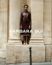 Model Saadia Bodenmann standing between limestone columns wearing a brown leather button-up shirt and coordinated midi skirt.