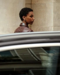 Profile of a woman with short natural hair wearing a brown leather jacket viewed over a car roof against a stone building wall.