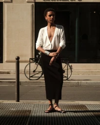 Model Saadia Bodenmann in a deep V-neck white top and black column skirt standing on a sunny street.