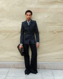 Model standing outdoors wearing a dark belted utility jacket and trousers while holding a black clutch bag.