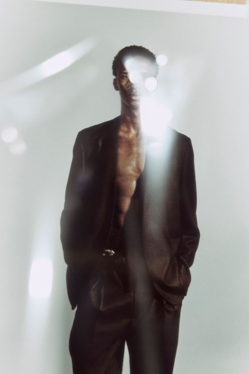 Model Baba Diagne poses shirtless in an open dark suit blazer and matching trousers against a light backdrop with foreground lens flares.