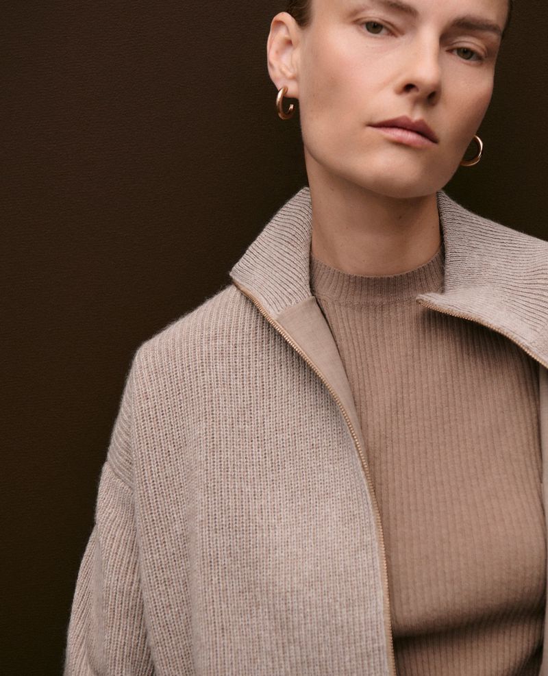 Model Ania Kisiel wearing layered taupe knitwear and gold hoop earrings against a dark brown studio background.