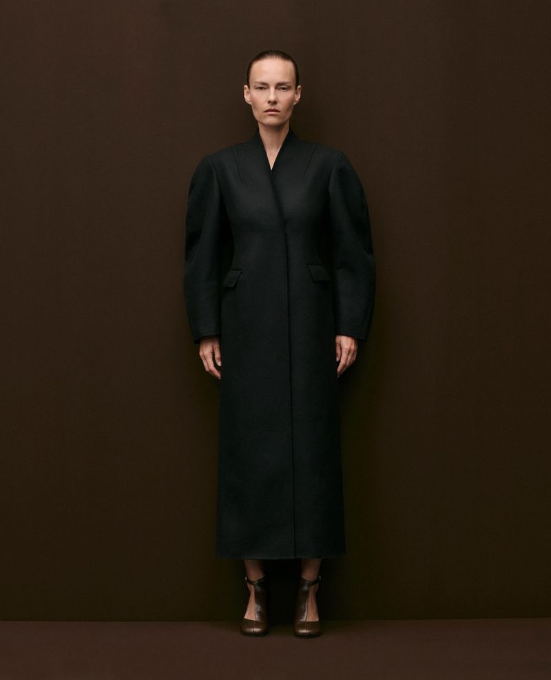 Model Ania Kisiel posing in a structured black ankle-length coat and dark heels against a solid dark brown backdrop.