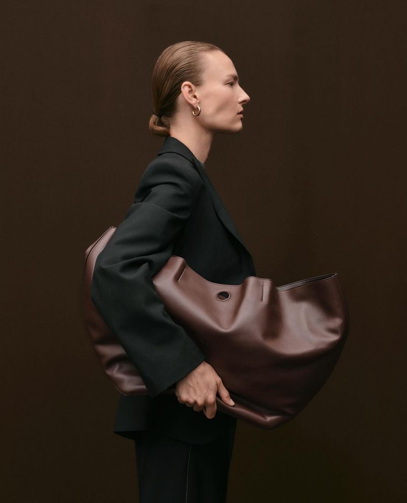Profile shot of model wearing a black blazer and carrying a large dark brown leather shoulder bag against a solid dark brown background.