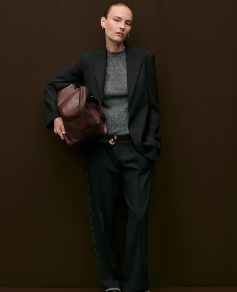 Model Ania Kisiel posing in a dark tailored trouser suit, grey crewneck, and brown leather bag against a dark brown background.