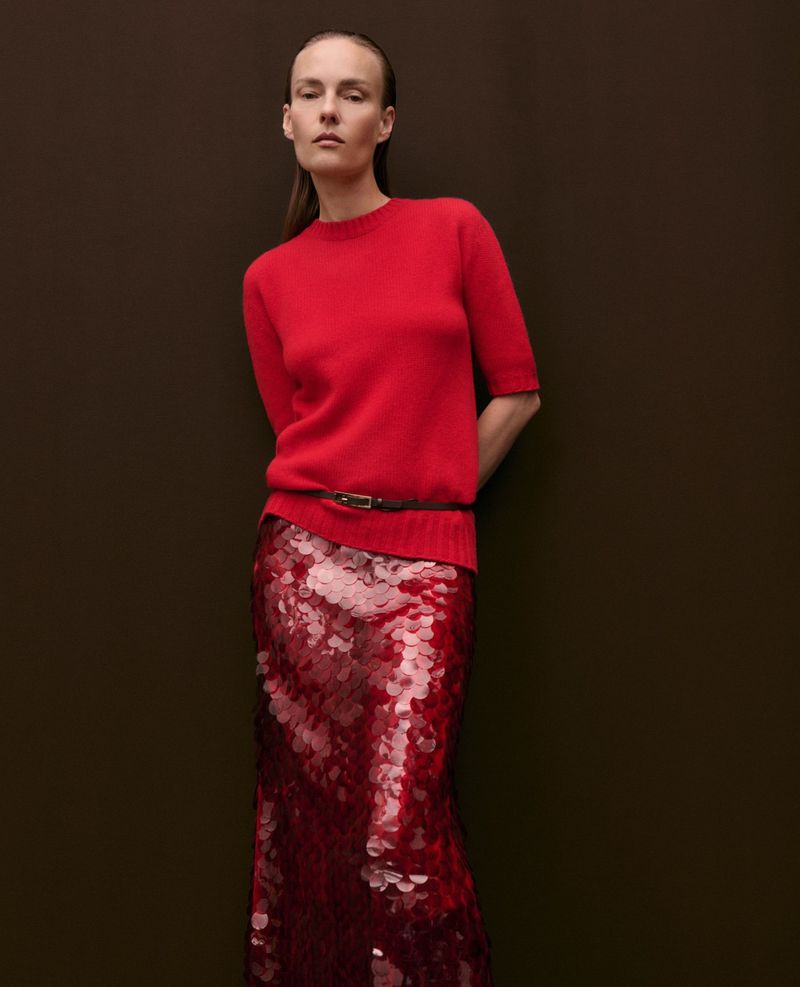 Model Ania Kisiel in a red short-sleeve knit sweater and red sequin maxi skirt against a dark brown studio backdrop.
