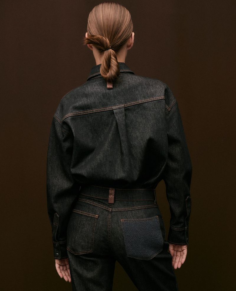 Model Ania Kisiel seen from the back wearing a dark denim shirt and matching jeans with contrast stitching against a brown backdrop.